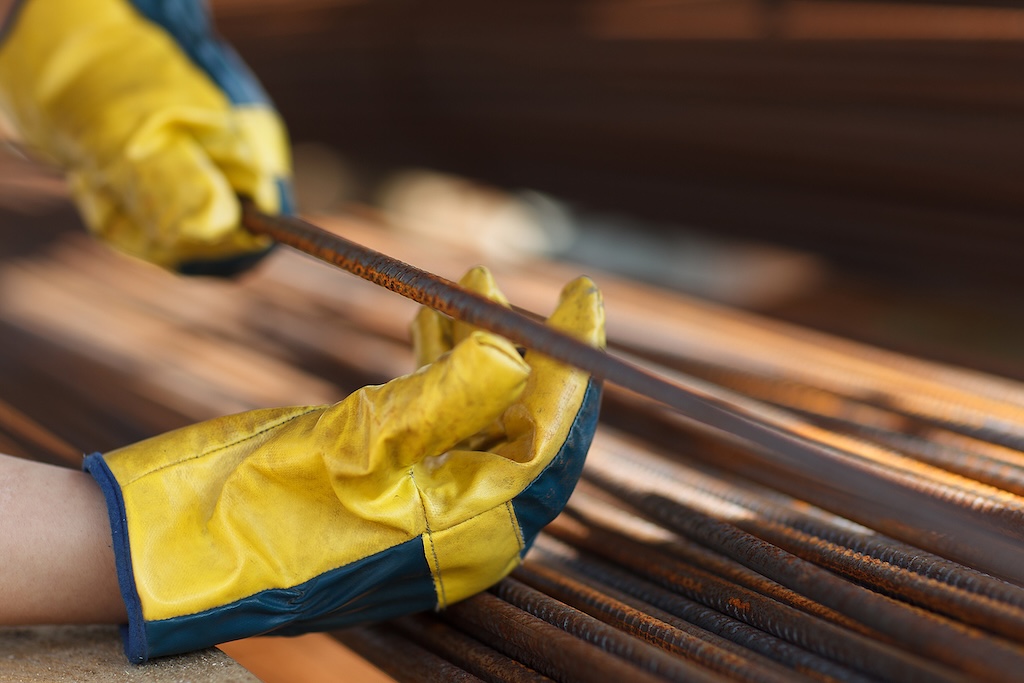 A woman in a yellow helmet and safety gear is working with metal bars. She is bending over to pick up a bar and is wearing gloves